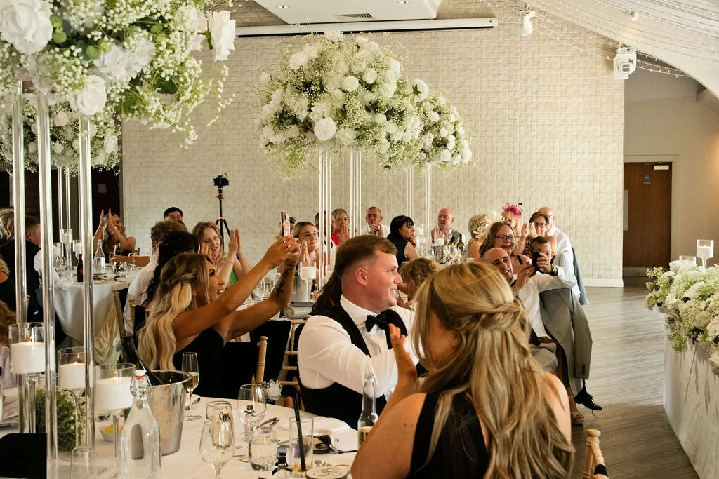 Wedding guests enjoy speeches at a reception table decorated with tall white floral centrepieces and glowing candles.