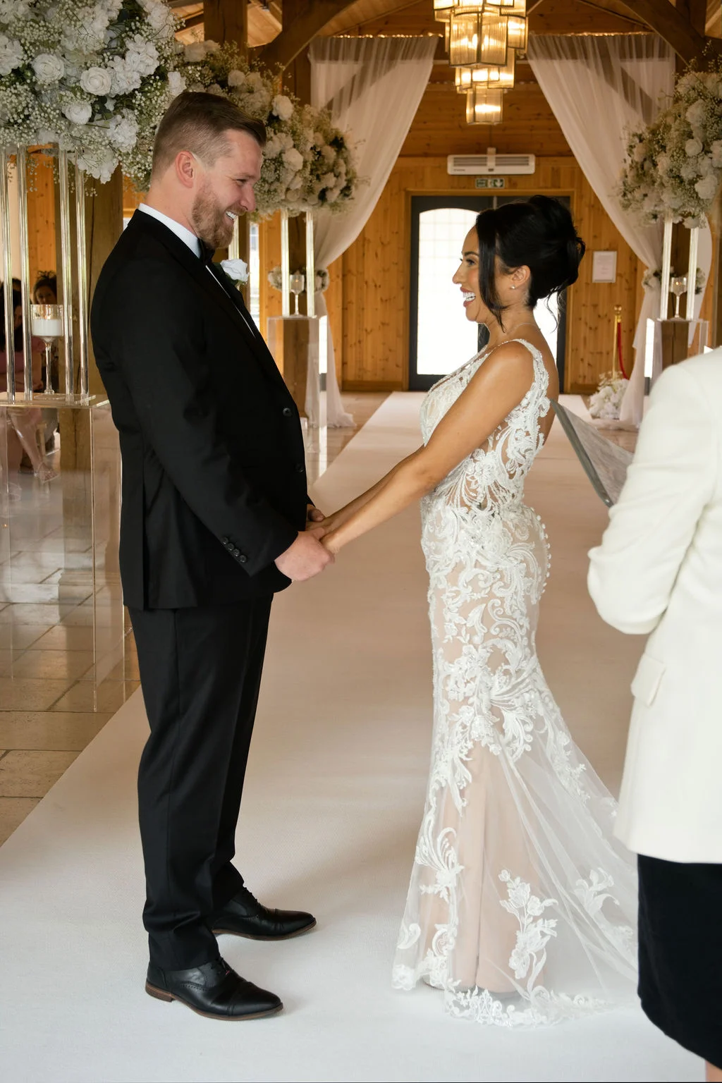 A smiling bride and groom hold hands facing each other during their civil ceremony inside a light-filled oak framed barn.