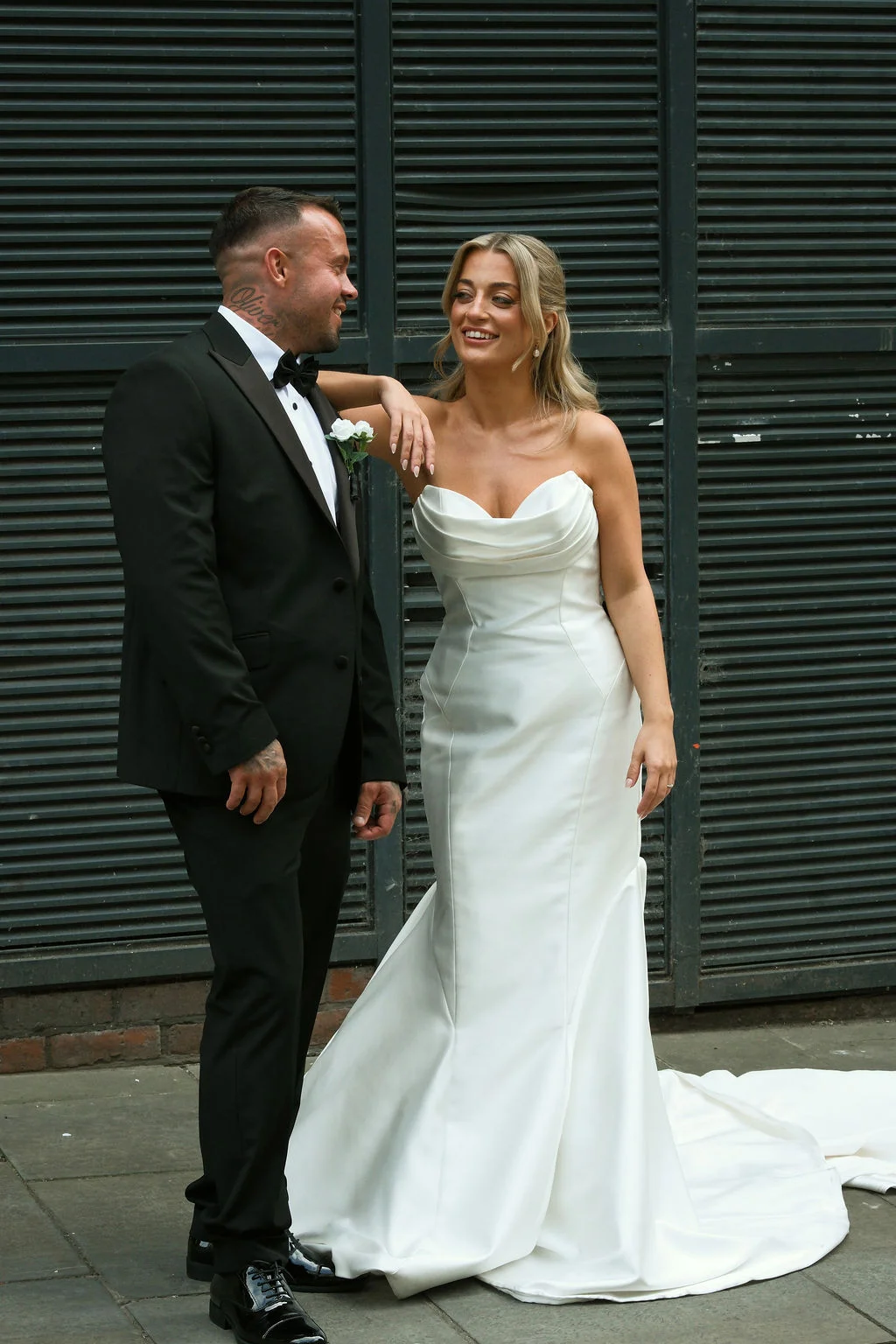 A newlywed couple shares a joyful glance outside against a dark slatted background, with the bride wearing an elegant strapless satin gown and the groom in a classic black tuxedo.