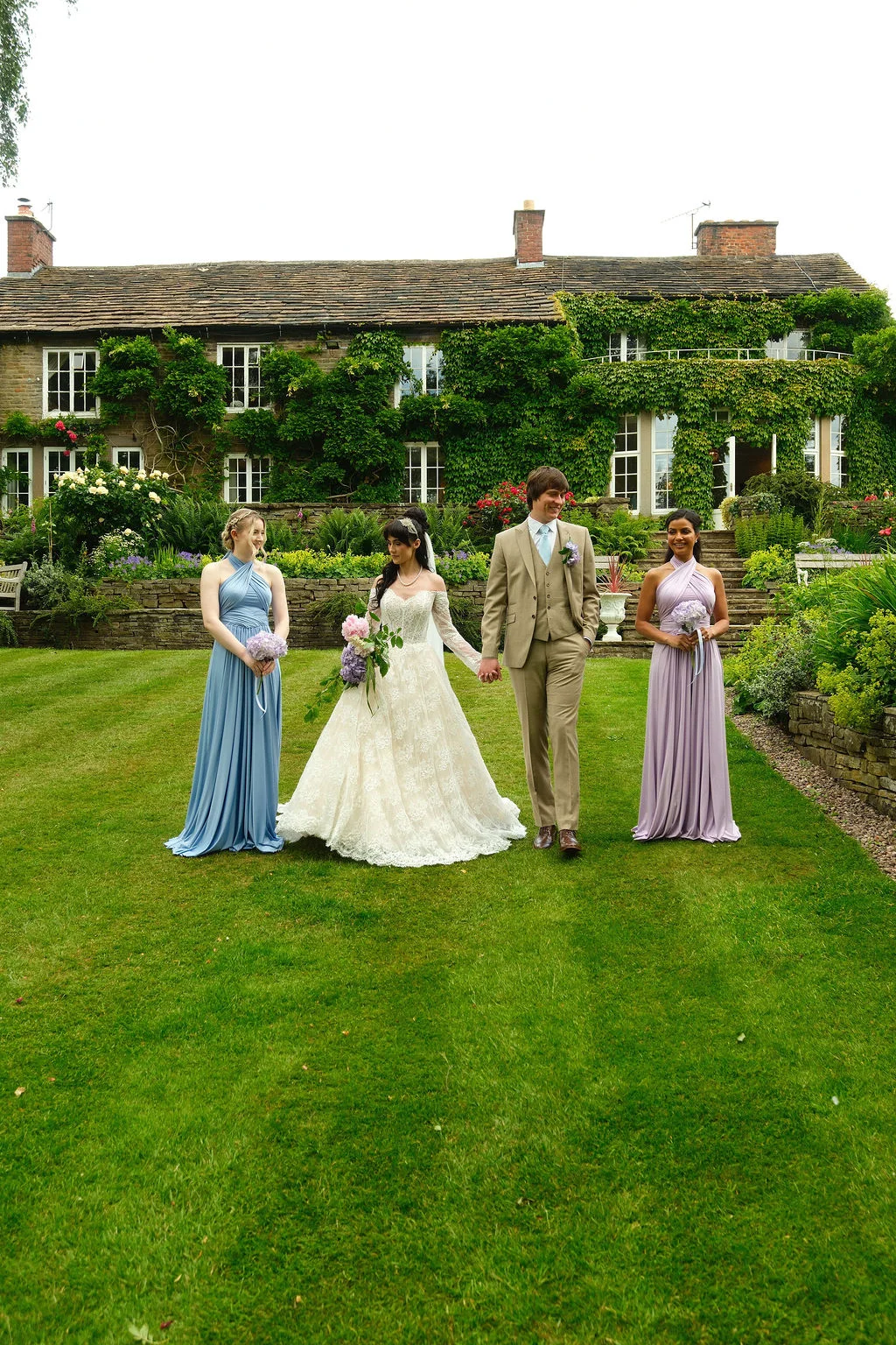 A bride in an off-the-shoulder lace gown and a groom in a tan suit walk across the lawn of a historic country house, flanked by two bridesmaids in pastel multi-way dresses.