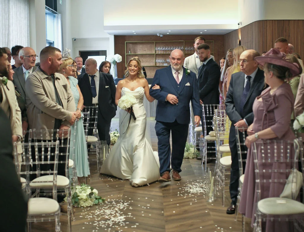 A smiling bride walks down the aisle arm in arm with her father, holding a white rose bouquet while guests look on during the wedding ceremony.