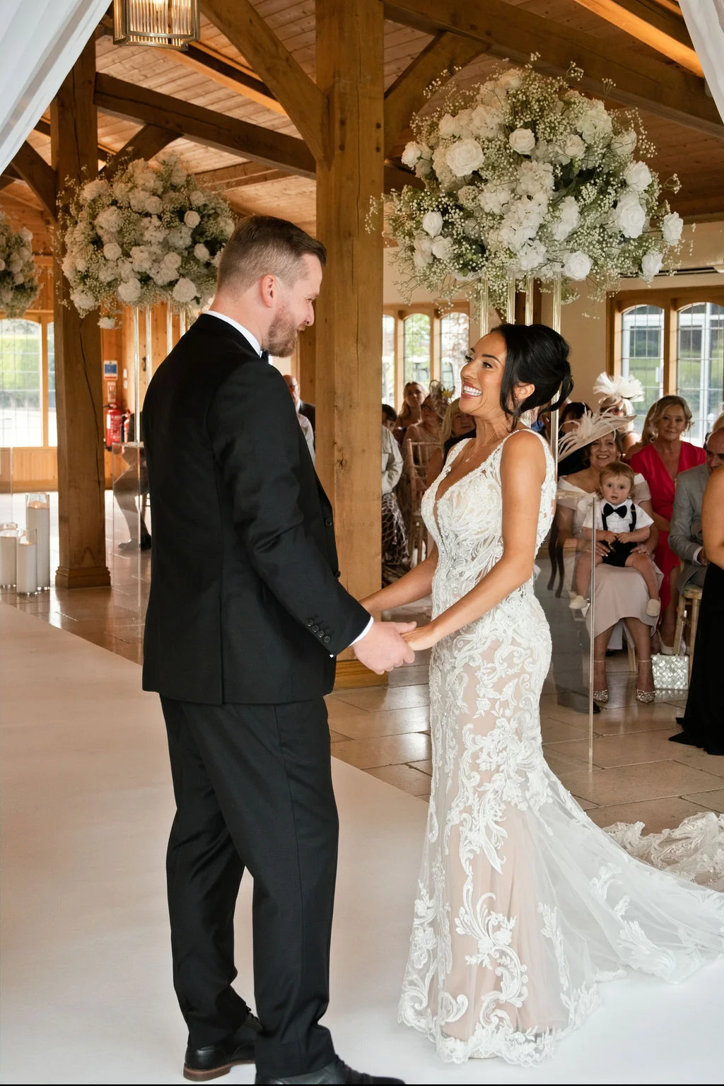 A joyful bride and groom hold hands and smile at each other during their indoor wedding ceremony inside an oak-framed barn decorated with white floral arrangements.