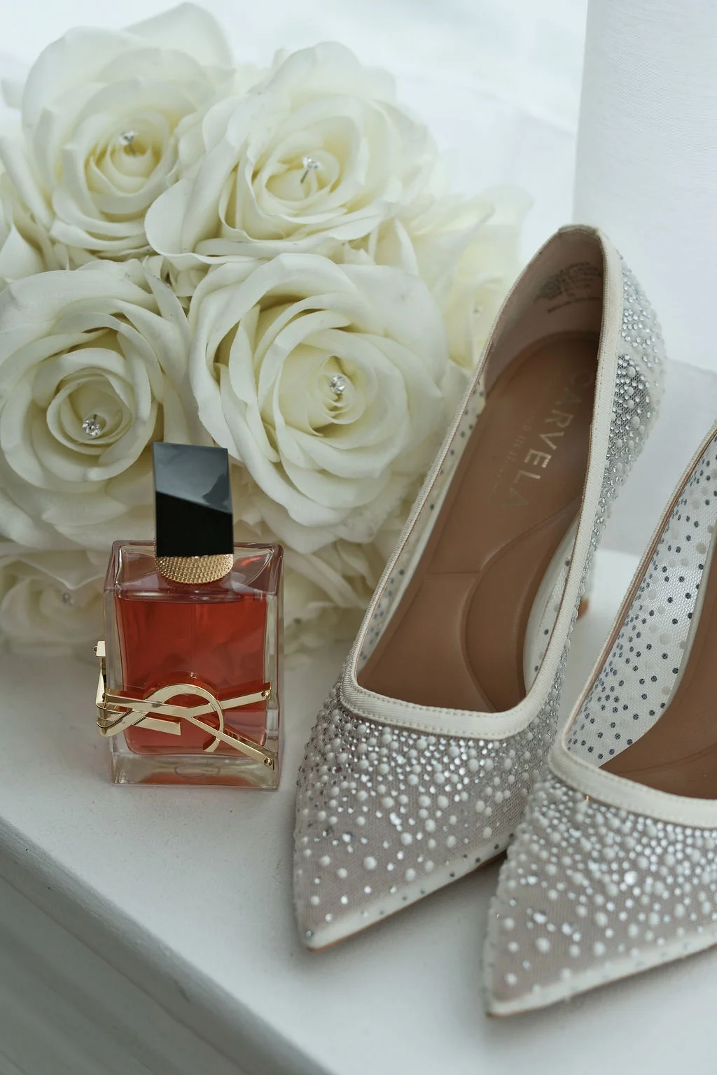 An elegant bridal flat lay features sparkling crystal heels, a square perfume bottle with a gold emblem, and a lush bouquet of white roses.