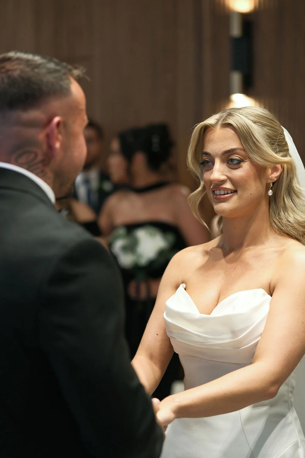 A glowing bride smiles lovingly at her groom during their wedding ceremony while holding hands.