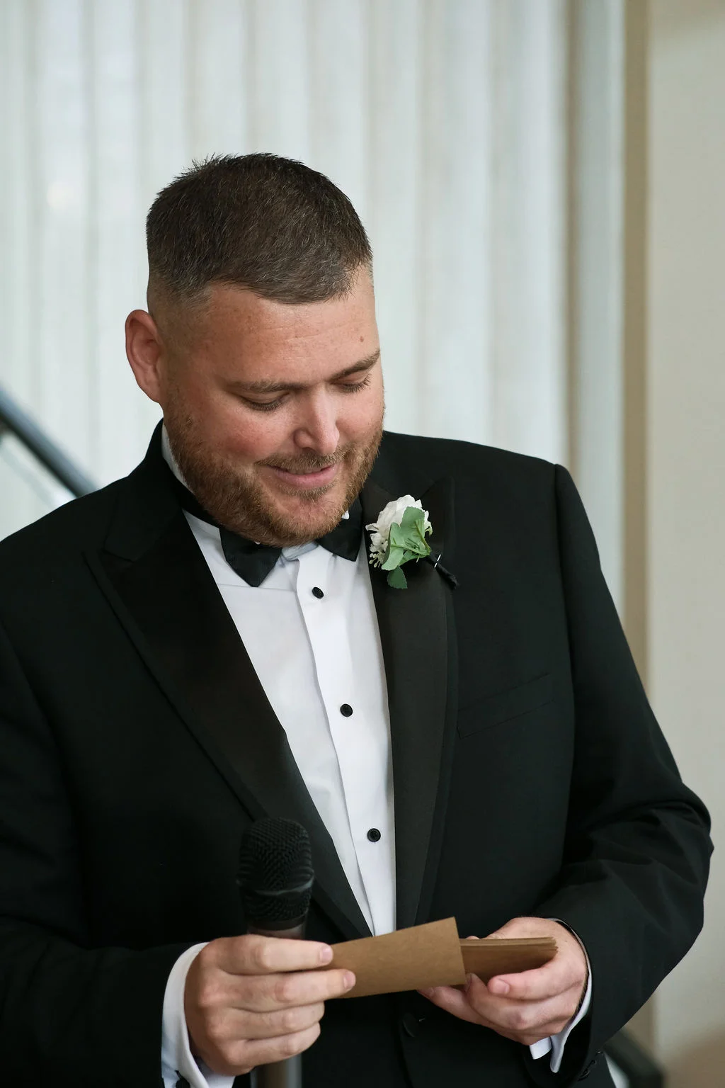 A groom in a black tuxedo and bow tie smiling down at speech notes while holding a microphone during the wedding reception.