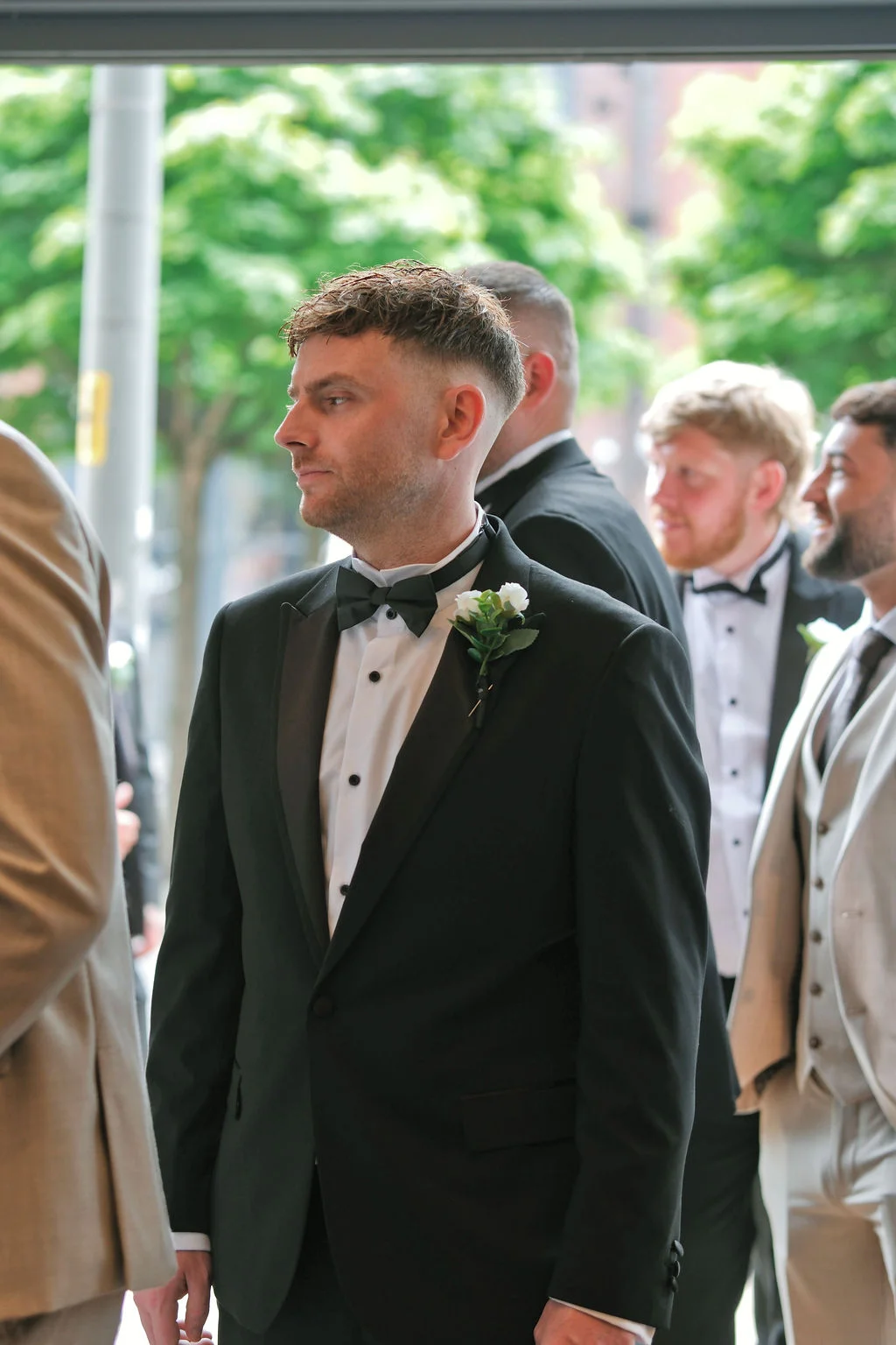A groom in a classic black tuxedo and bow tie waits attentively alongside his groomsmen during the wedding ceremony.