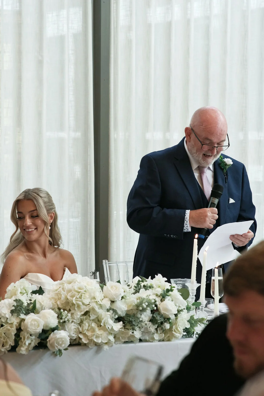A father of the bride delivers a speech into a microphone at the top table, while the smiling bride looks on beside a lush white floral arrangement.
