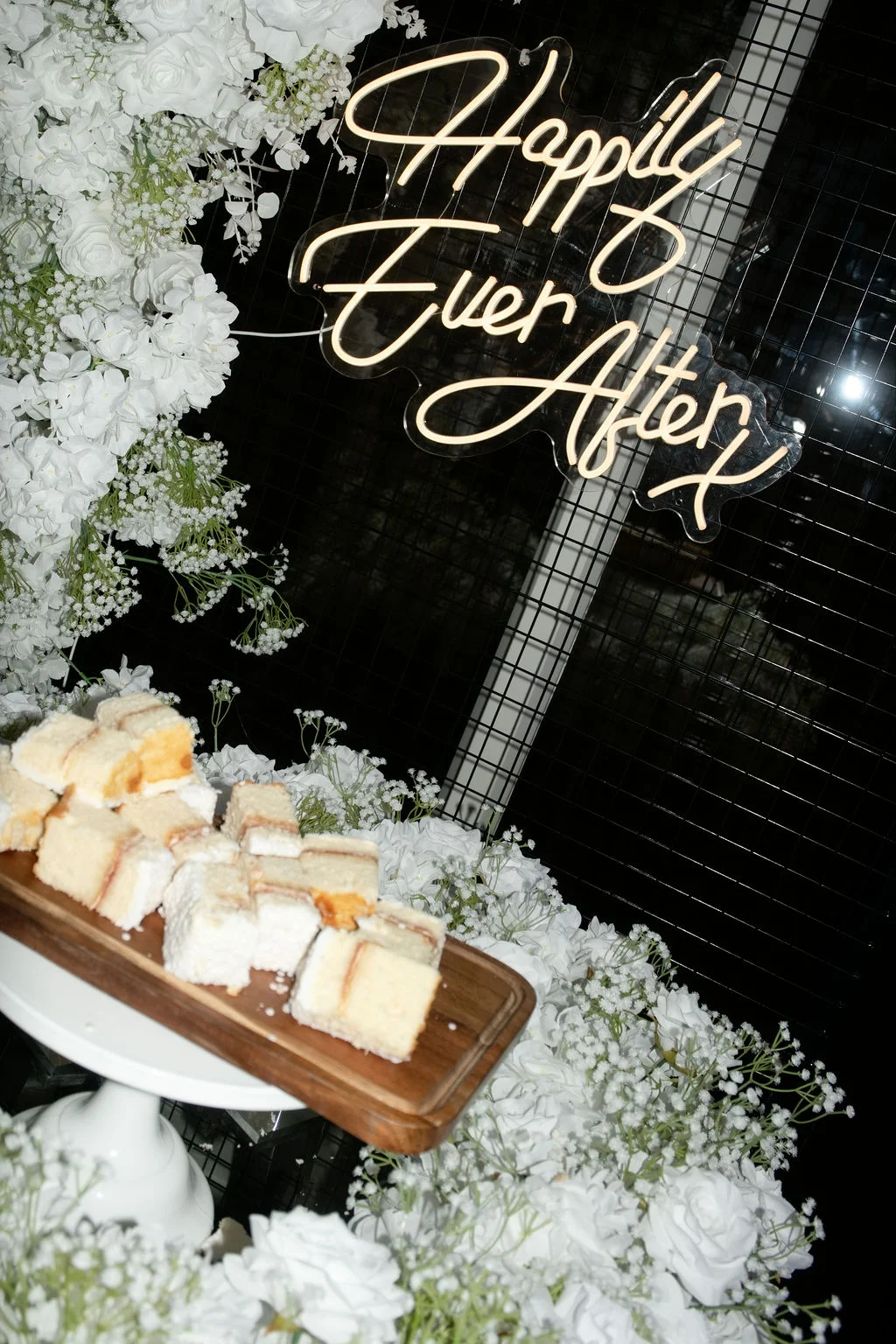 A close-up shot of a wedding dessert table featuring sponge cake slices on a wooden board, surrounded by cascading white roses and baby's breath beneath a glowing neon sign.