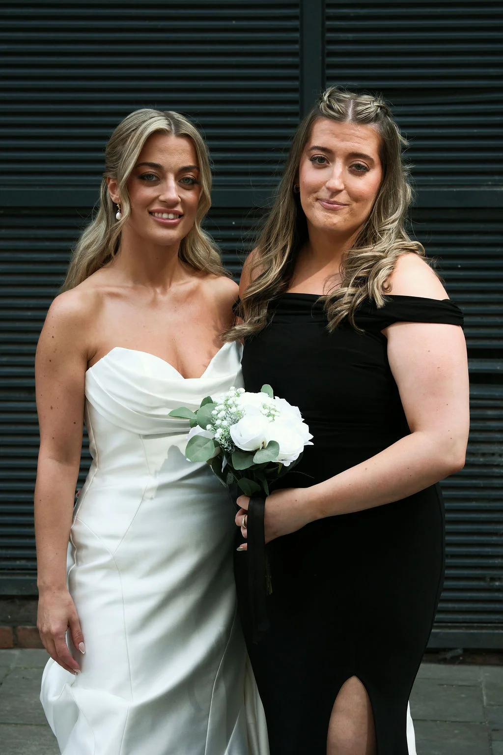 A smiling couple pose together outdoors against a dark slatted backdrop, one wearing a strapless white satin gown and the other in a black off-the-shoulder dress holding a bouquet of white roses.