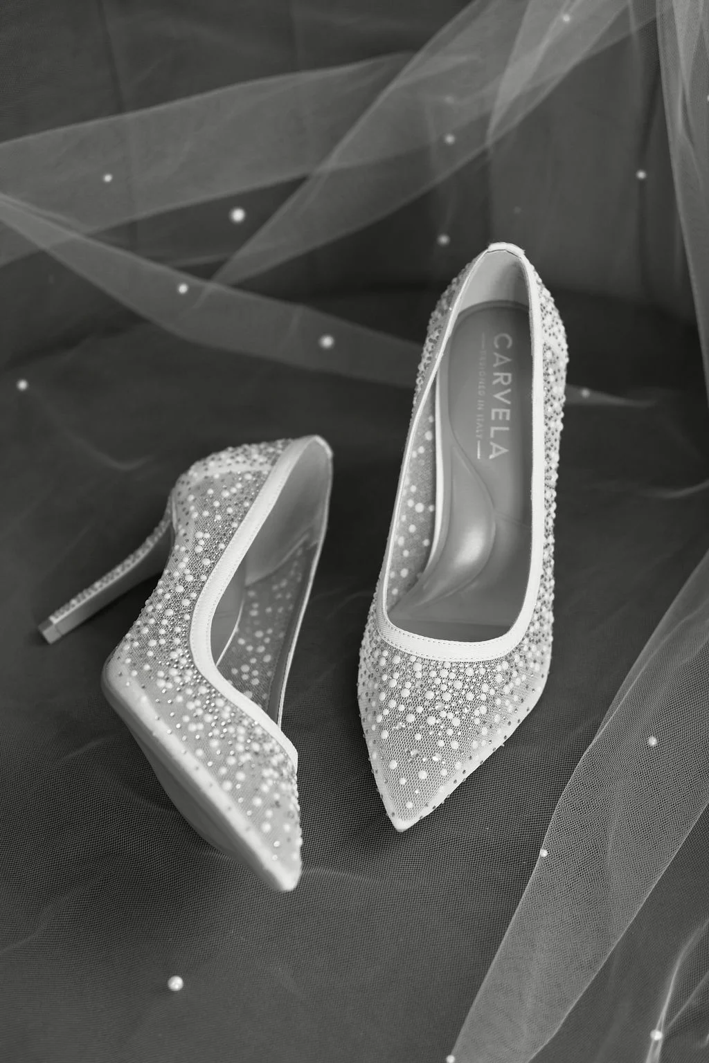 An elegant black and white flat lay features a pair of crystal and pearl-embellished bridal heels nestled beneath a delicate pearl-studded veil.