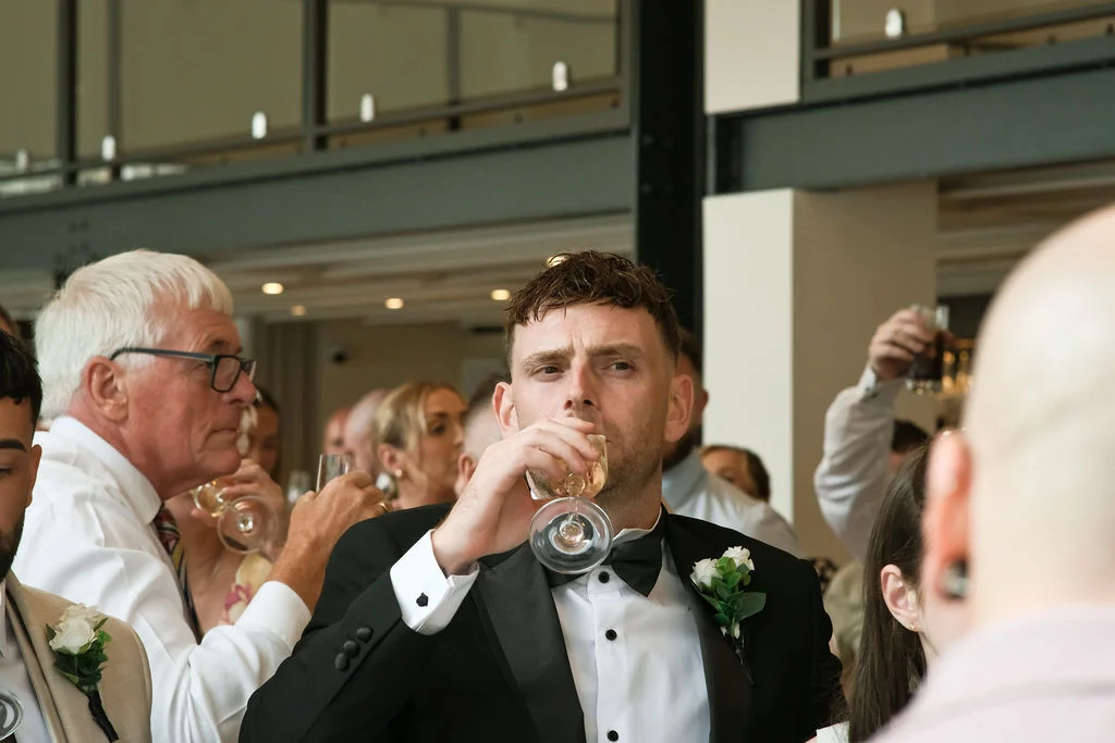 A groom in a black tuxedo and bow tie takes a drink from a glass during wedding speeches while surrounded by guests.