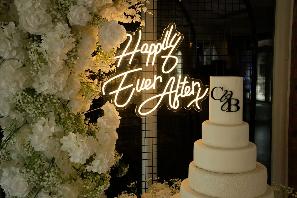 A stunning four-tier white wedding cake featuring a custom monogram sits next to a glowing neon sign and a lush white floral wall.