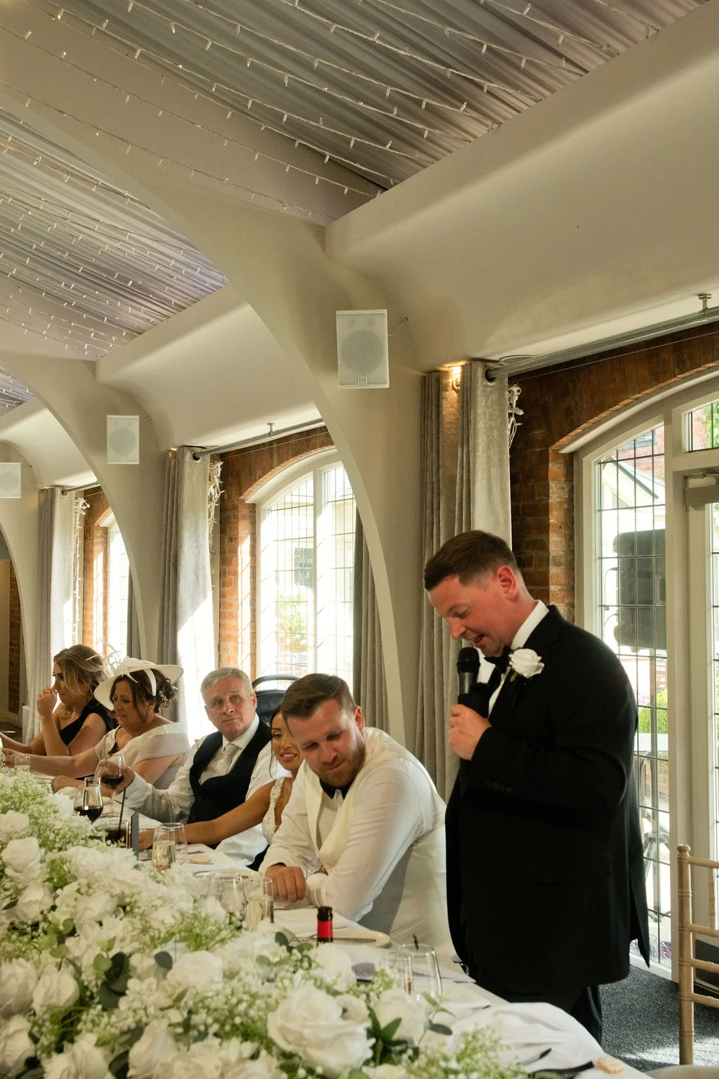 A groom in a black tuxedo delivers his wedding speech at the top table, adorned with lush white floral arrangements and framed by draped arches.