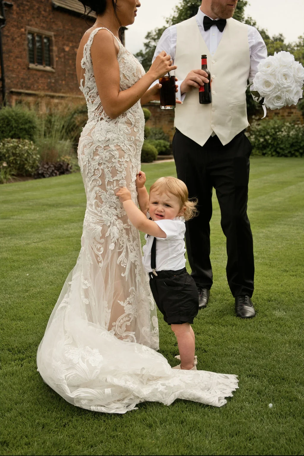 A distressed toddler in braces and shorts clutches the ornate lace gown of a bride standing on a manicured lawn next to a groom holding drinks.