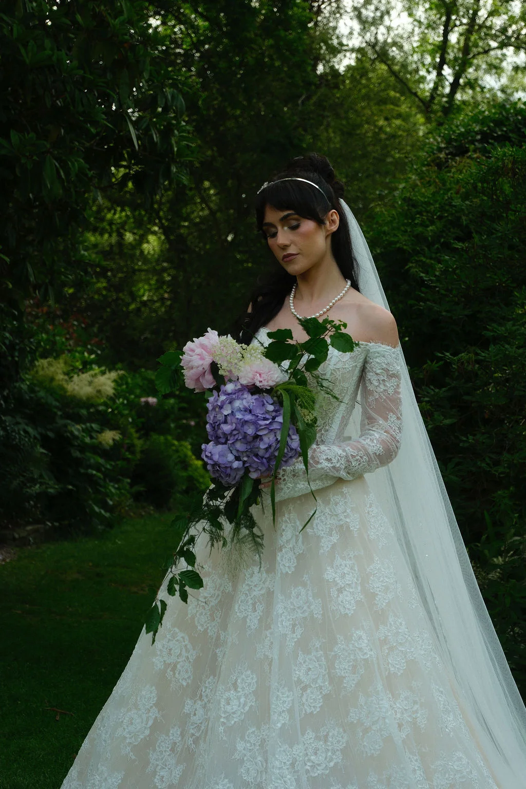 A stunning bride stands in a lush woodland holding a cascading bouquet of purple hydrangeas and pink peonies, wearing an off-the-shoulder lace gown and a chapel veil.