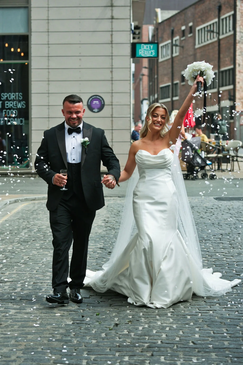 A newlywed couple walk hand in hand along a cobbled city street as white confetti falls around them, with the bride raising her bouquet in celebration.