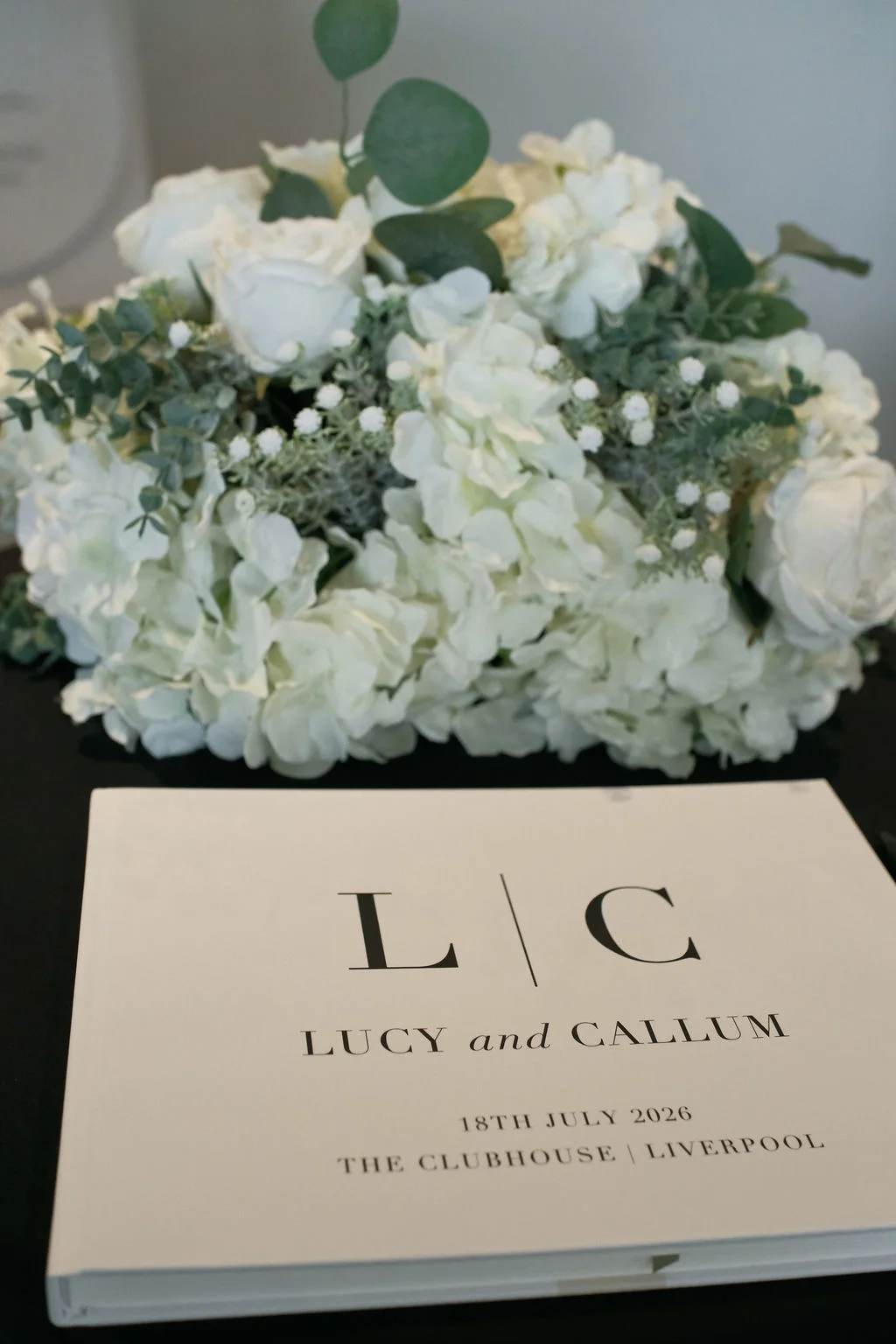 A close-up shot of a modern minimalist wedding guest book featuring a black monogram and text, resting below a lush arrangement of white roses, hydrangeas, and eucalyptus.