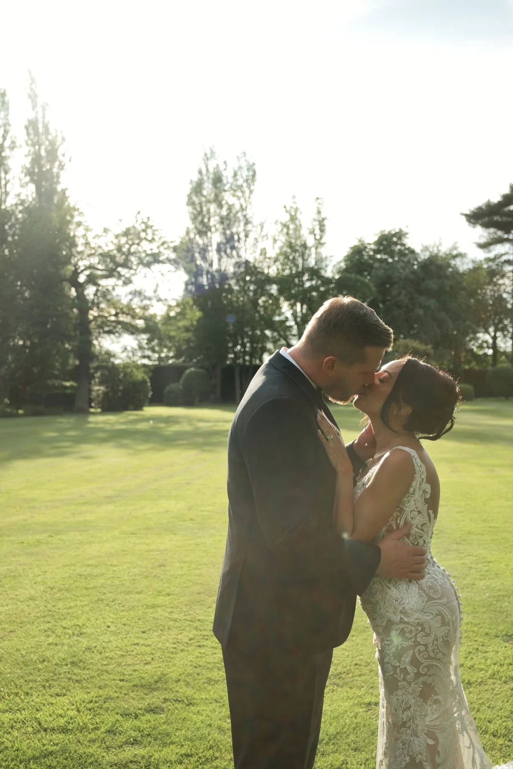 A newlywed couple shares a romantic kiss in a sunlit manicured garden during golden hour, with the bride wearing an intricate lace gown.