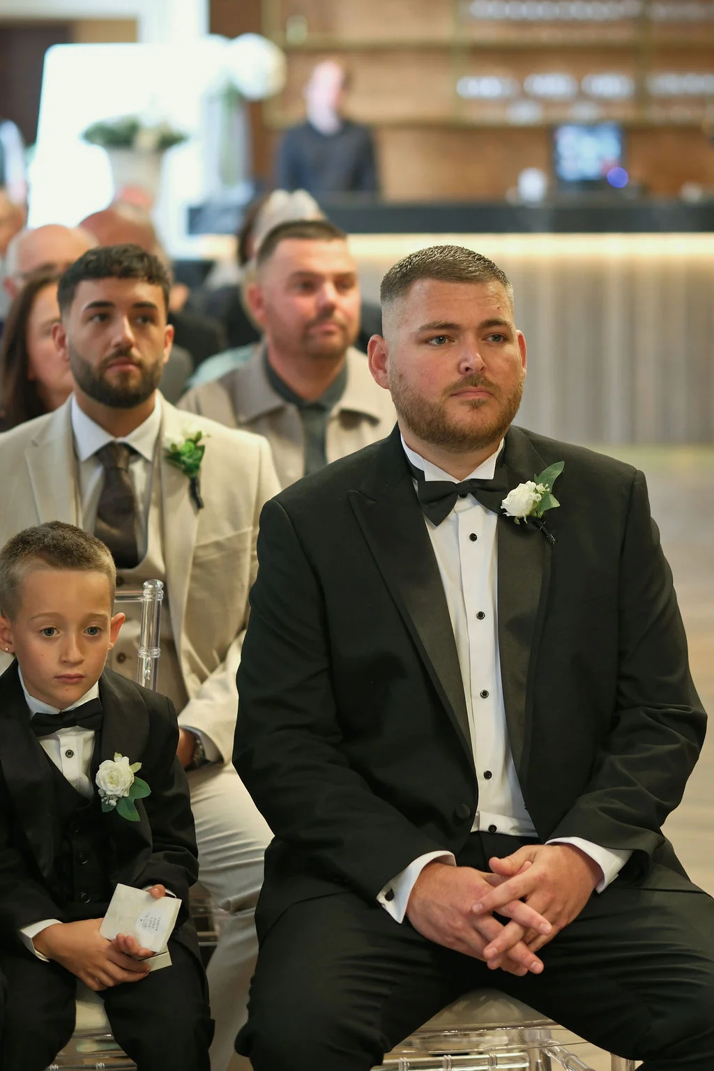 A groom in a classic black tuxedo with a white buttonhole sits attentively at the ceremony alongside his groomsmen and page boy.