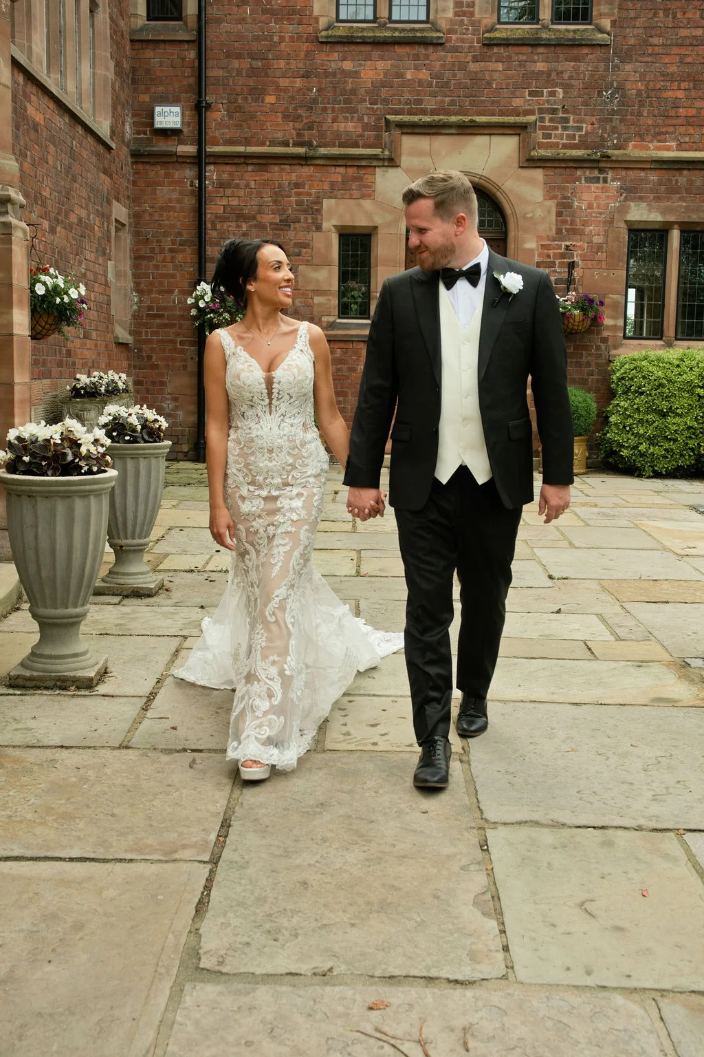 A newlywed couple happily walks hand-in-hand across a stone courtyard outside a red brick manor house, sharing a loving glance.