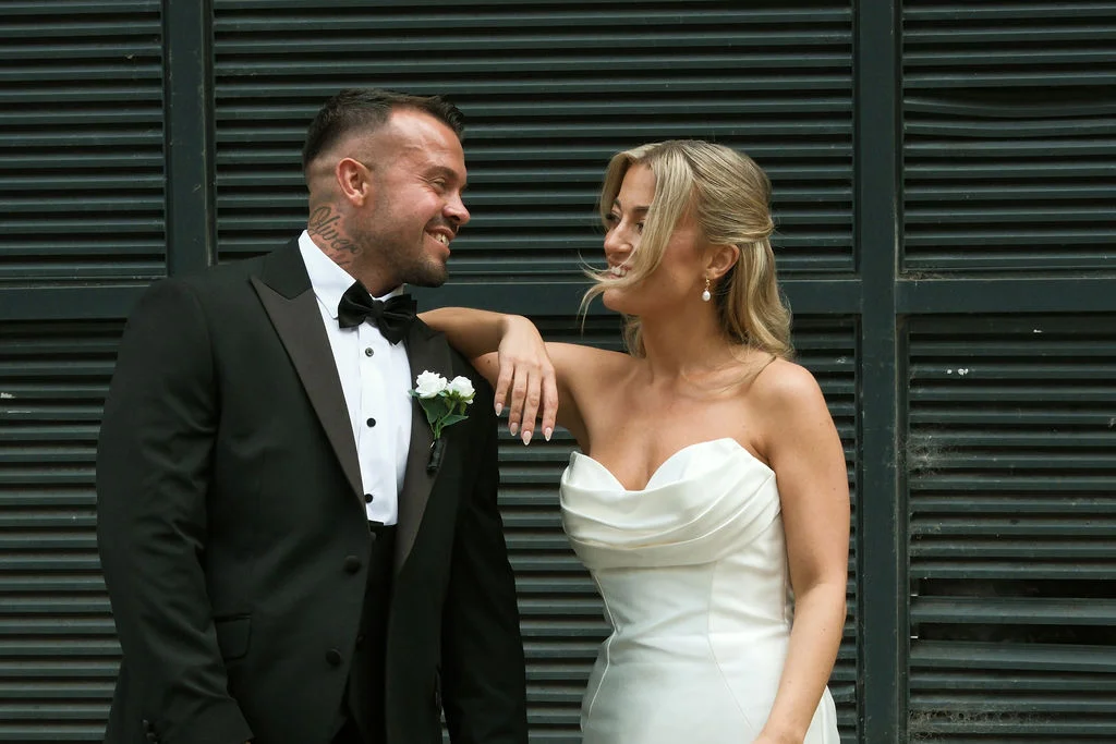 A smiling bride leaning against her groom in a black tuxedo while standing in front of a dark slatted background.