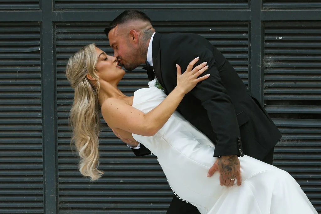 A groom dips and kisses his bride, who wears a strapless satin gown with button details and has long blonde hair in a half-up style.