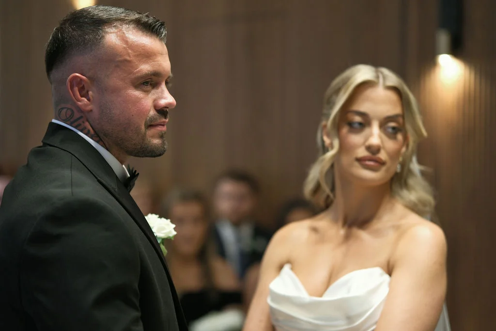 A groom in a black tuxedo and the bride in a strapless white satin gown exchange a tender glance during their indoor ceremony.