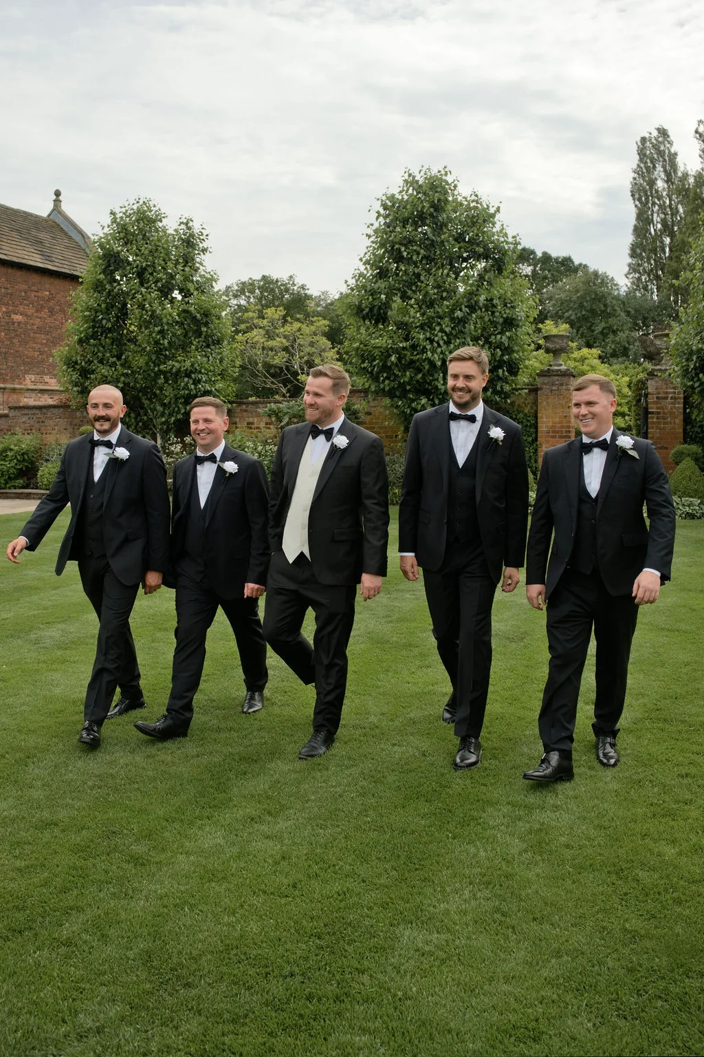 Five groomsmen in classic black tuxedos and bow ties walk together across a manicured lawn at a country house wedding venue.