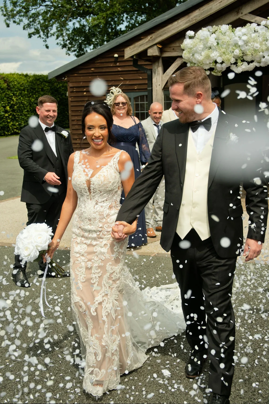 A joyful newlywed couple walks hand in hand out of a wooden barn while guests throw white confetti around them.