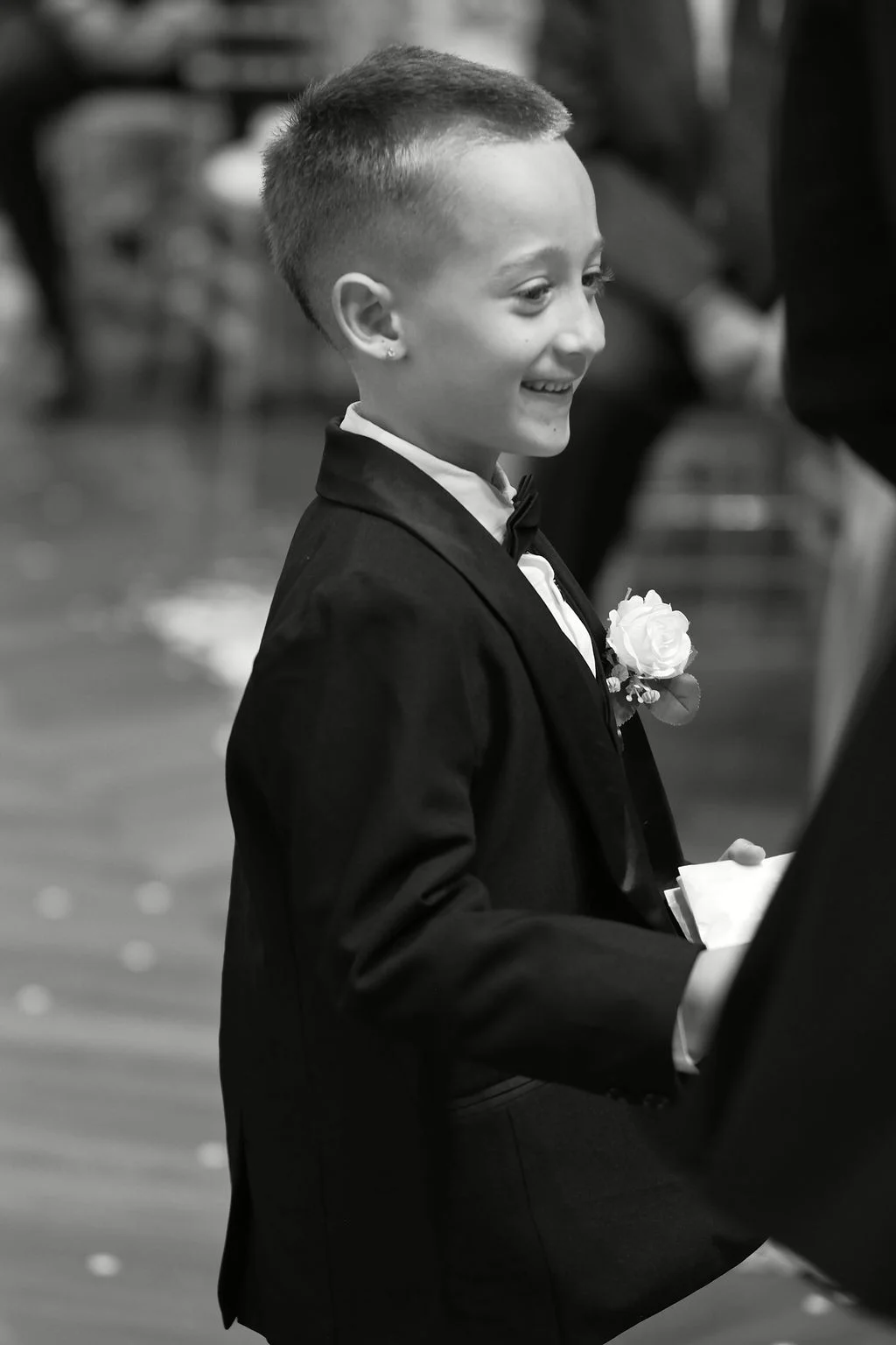 A young page boy in a black tuxedo and bow tie smiles during the wedding ceremony.