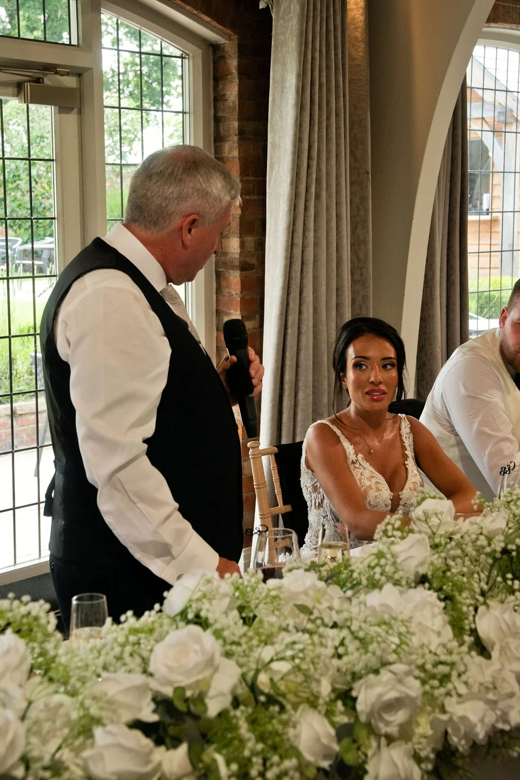 The father of the bride delivers a speech at the top table while the bride listens attentively beside a lush arrangement of white roses.