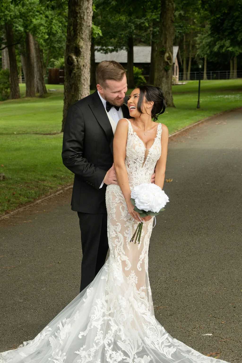 A newlywed couple shares a joyful, laughing glance while standing on a paved driveway surrounded by lush green trees and manicured lawns.