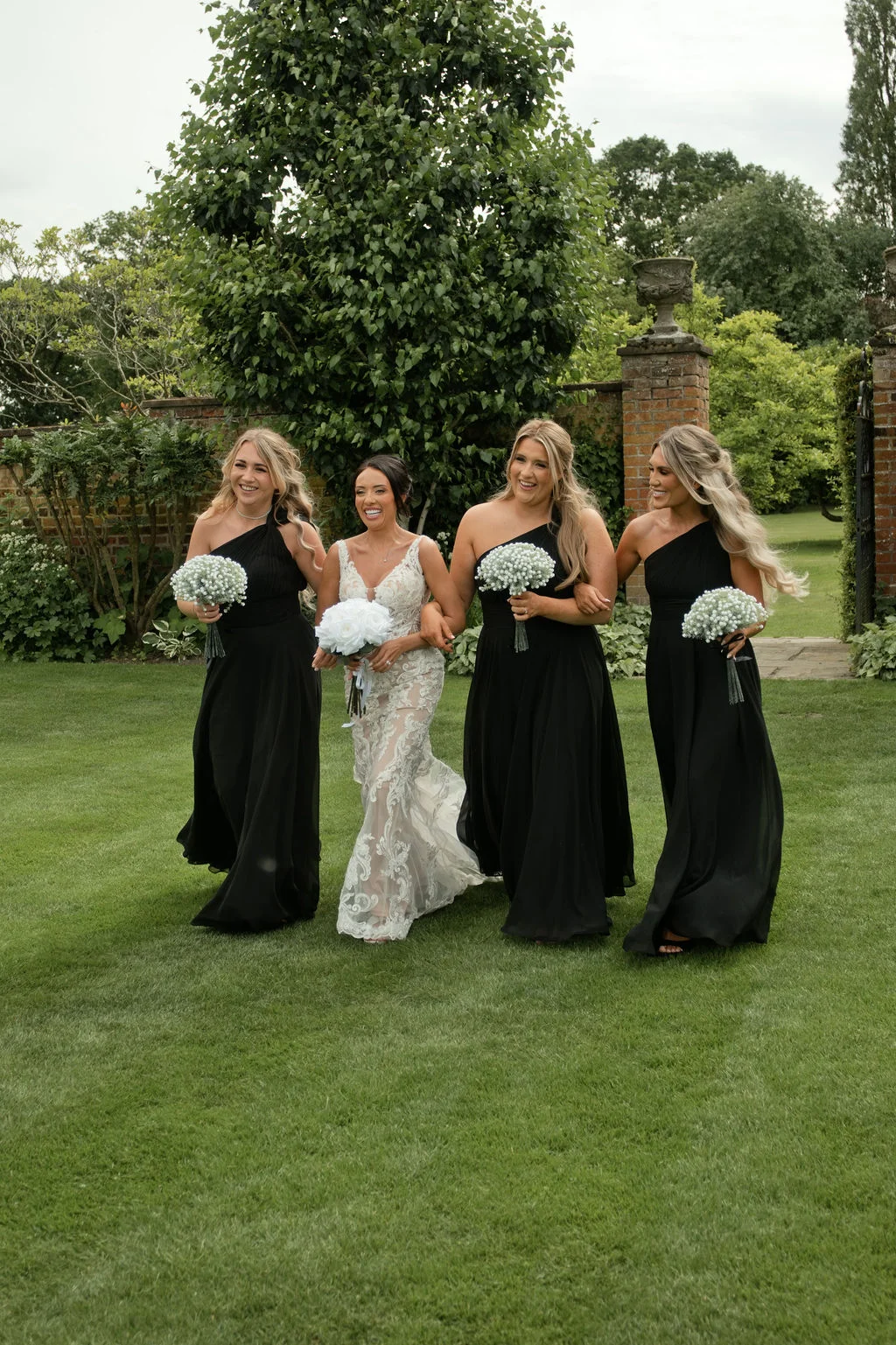 A laughing bride in a lace wedding dress and her four bridesmaids in matching black one-shoulder gowns walk across a lush green lawn, holding bouquets of white flowers.