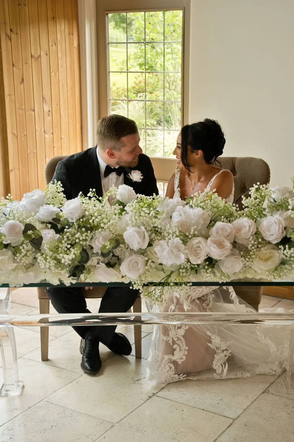 A newlywed bride and groom lovingly gaze at each other while seated at a glass signing table adorned with an abundant floral arrangement of white roses and gypsophila.