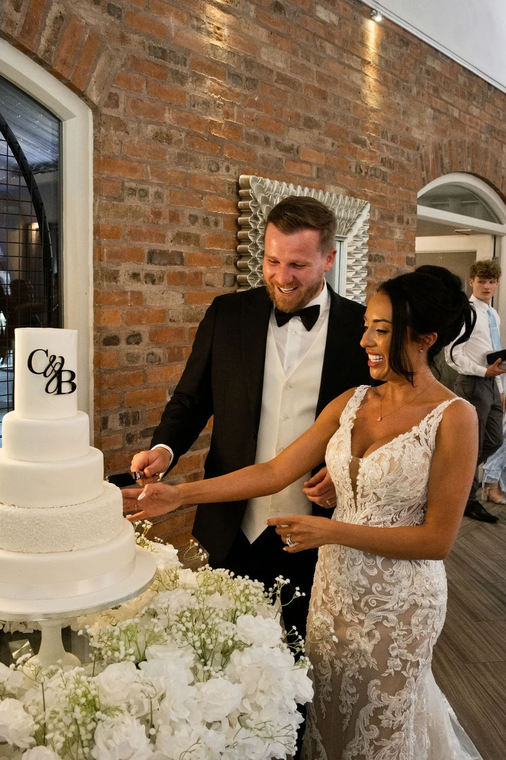 A smiling bride and groom cut into a white four-tiered wedding cake adorned with a monogram topper against an exposed brick wall backdrop.