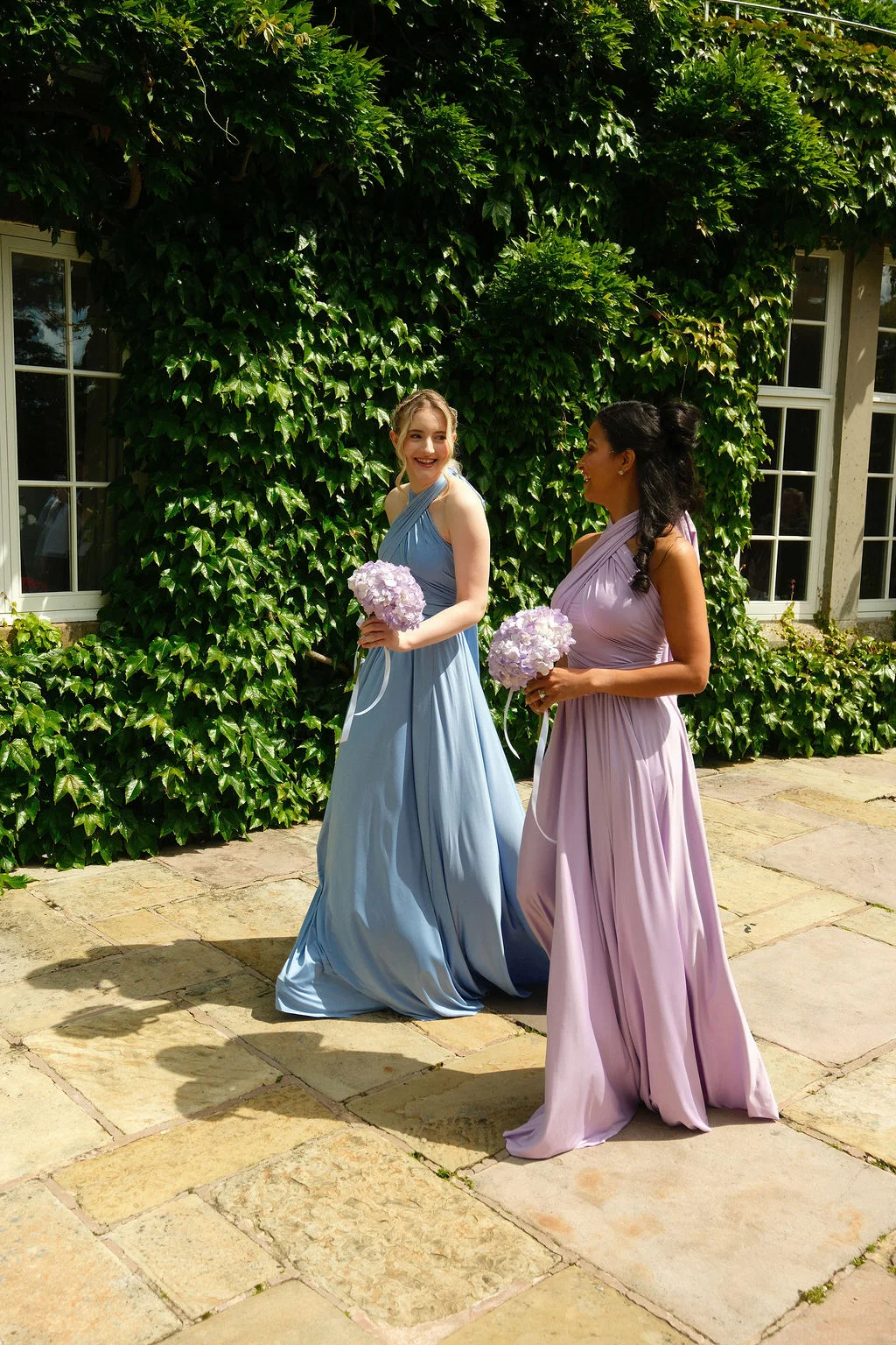 Two smiling bridesmaids in pastel blue and lilac halterneck dresses walk together on a stone patio in front of an ivy-covered wall, holding matching round bouquets.