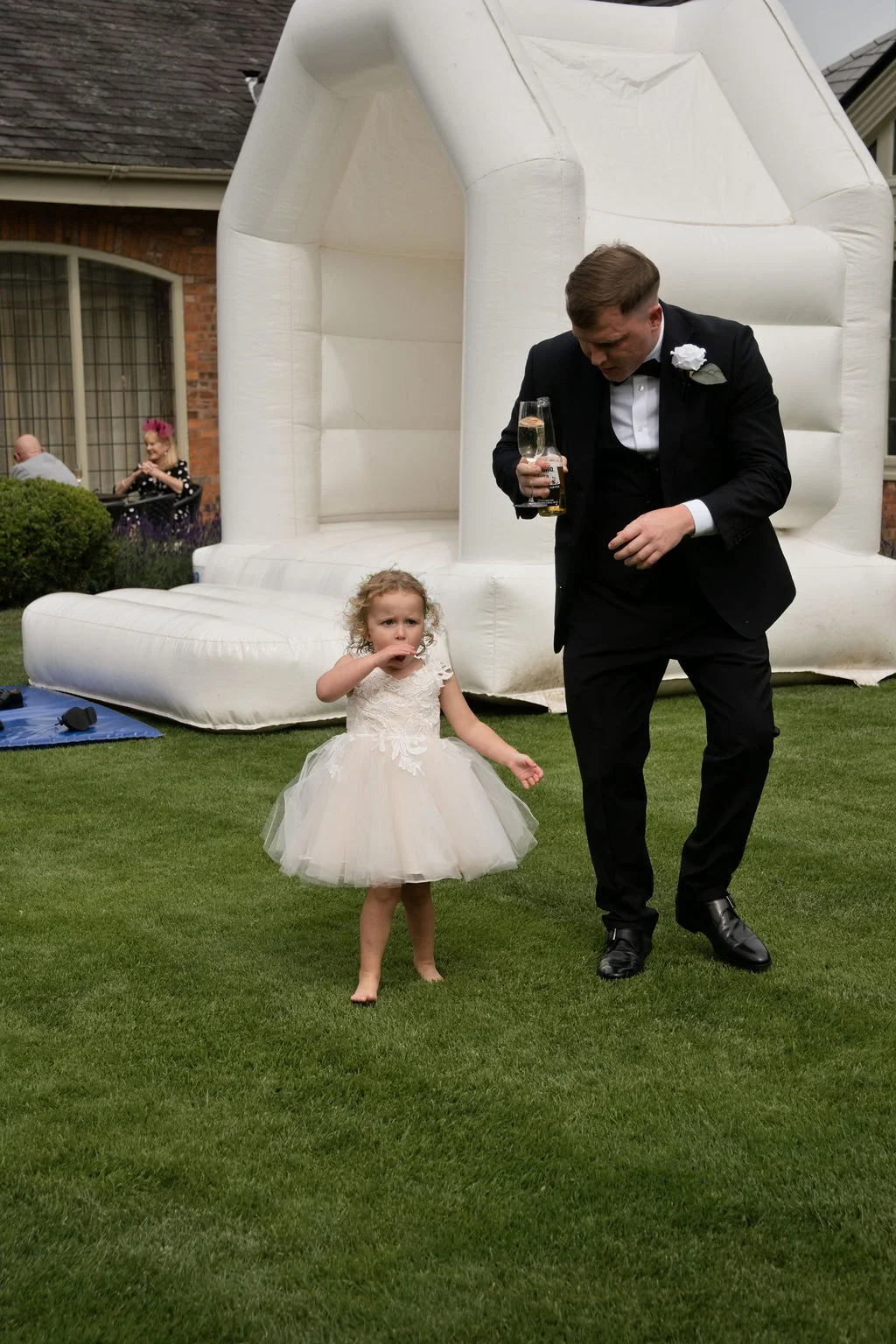 A young flower girl in a tulle dress walks barefoot on the lawn alongside a groom holding a drink in front of a white bouncy castle.
