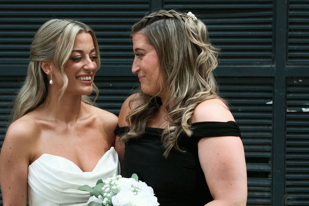 A smiling bride in a white gown shares a tender moment with a bridesmaid against a dark slatted background, holding a white floral bouquet.