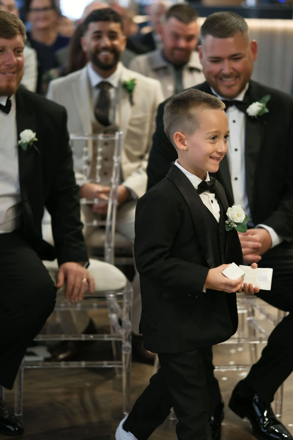 A young page boy happily carrying a ring box down the aisle while smiling guests look on during the wedding ceremony.