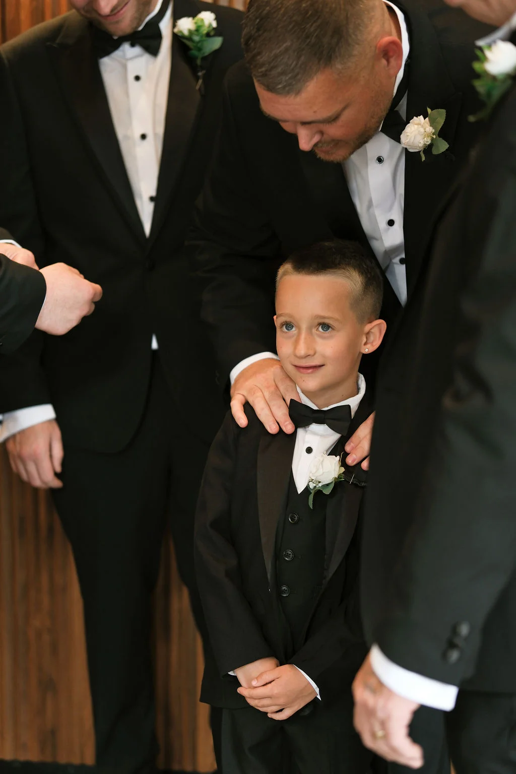 A young page boy wearing a matching black tuxedo and bow tie stands proudly while a groomsman gently places a hand on his shoulder.