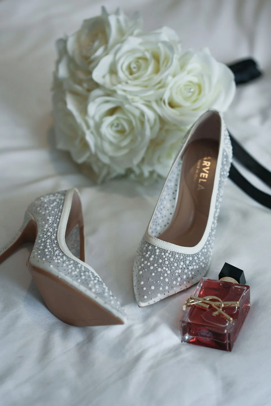 A bridal prep flat lay features embellished high heel shoes alongside a bottle of perfume and a white rose bouquet.
