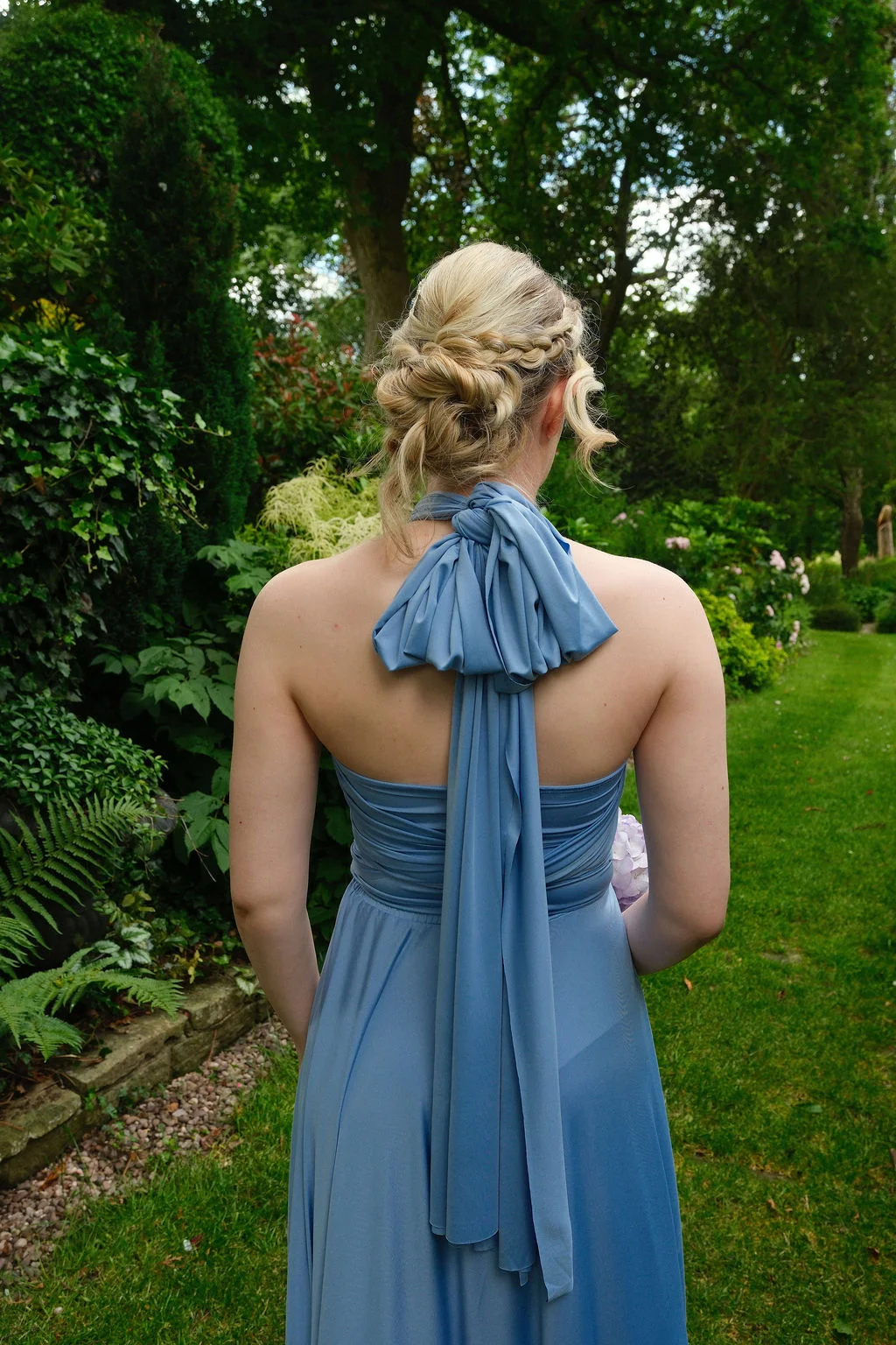A bridesmaid viewed from behind in a lush garden setting, showcasing an intricate braided low bun and a blue halterneck dress with a prominent fabric bow tied at the neck.