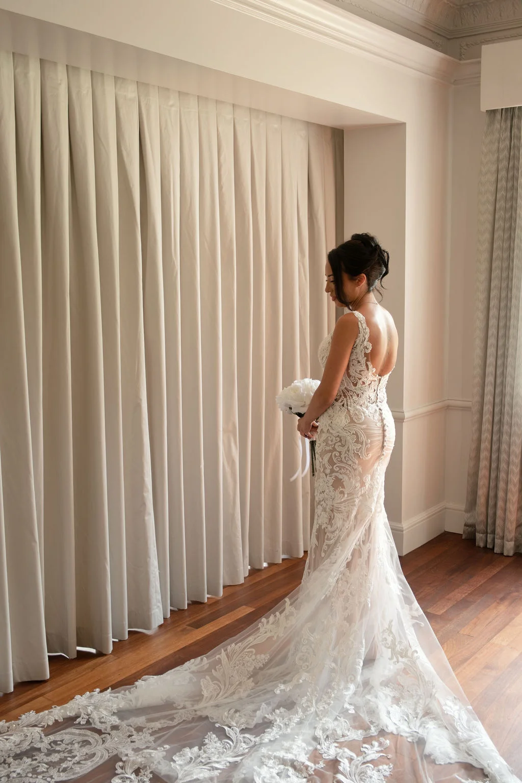 A bride standing near a large draped window wearing an intricate lace wedding dress with an illusion back and a long train, holding a white bouquet.