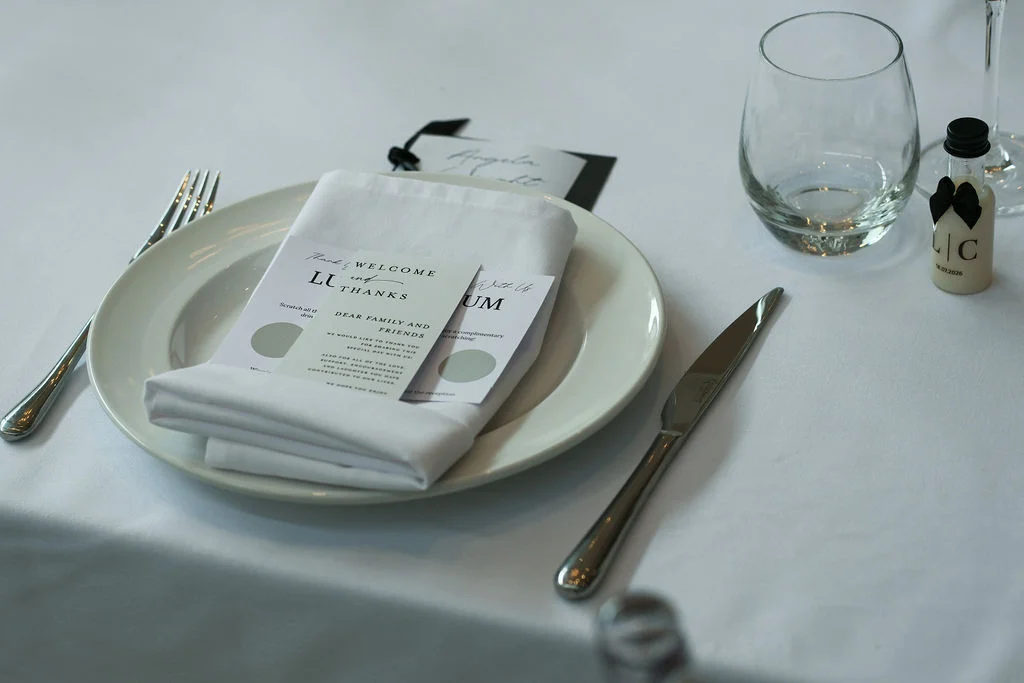 An elegantly set wedding table features a white plate topped with a folded napkin, printed cards, and a miniature favour bottle wearing a tiny black bow tie.