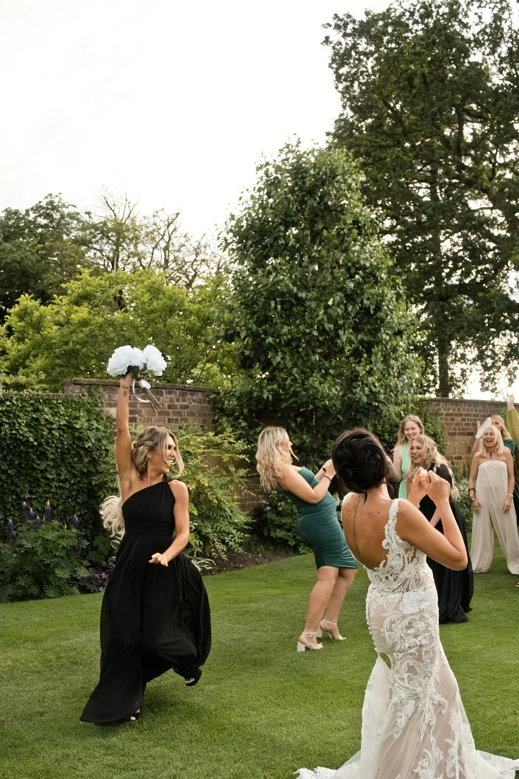 A smiling bride in a lace gown watches excitedly as a guest in a black dress catches the bouquet during an outdoor garden wedding celebration.