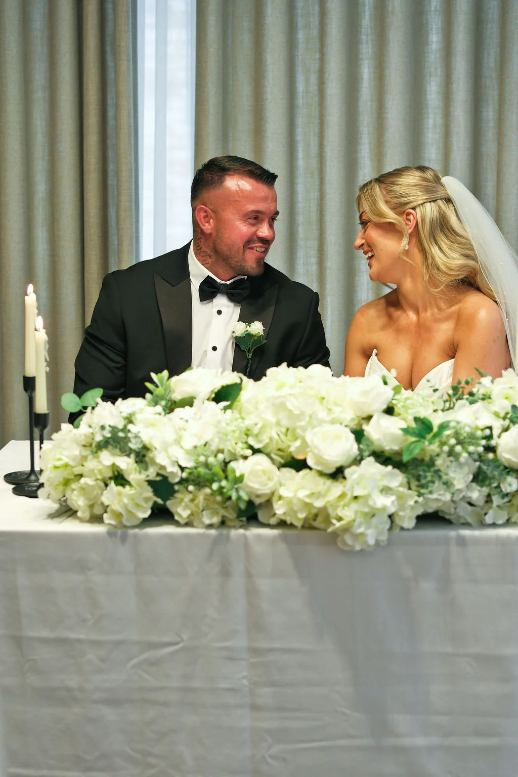 A newlywed couple shares a joyful moment seated at a white-linened top table adorned with a lush arrangement of white roses and glowing taper candles.