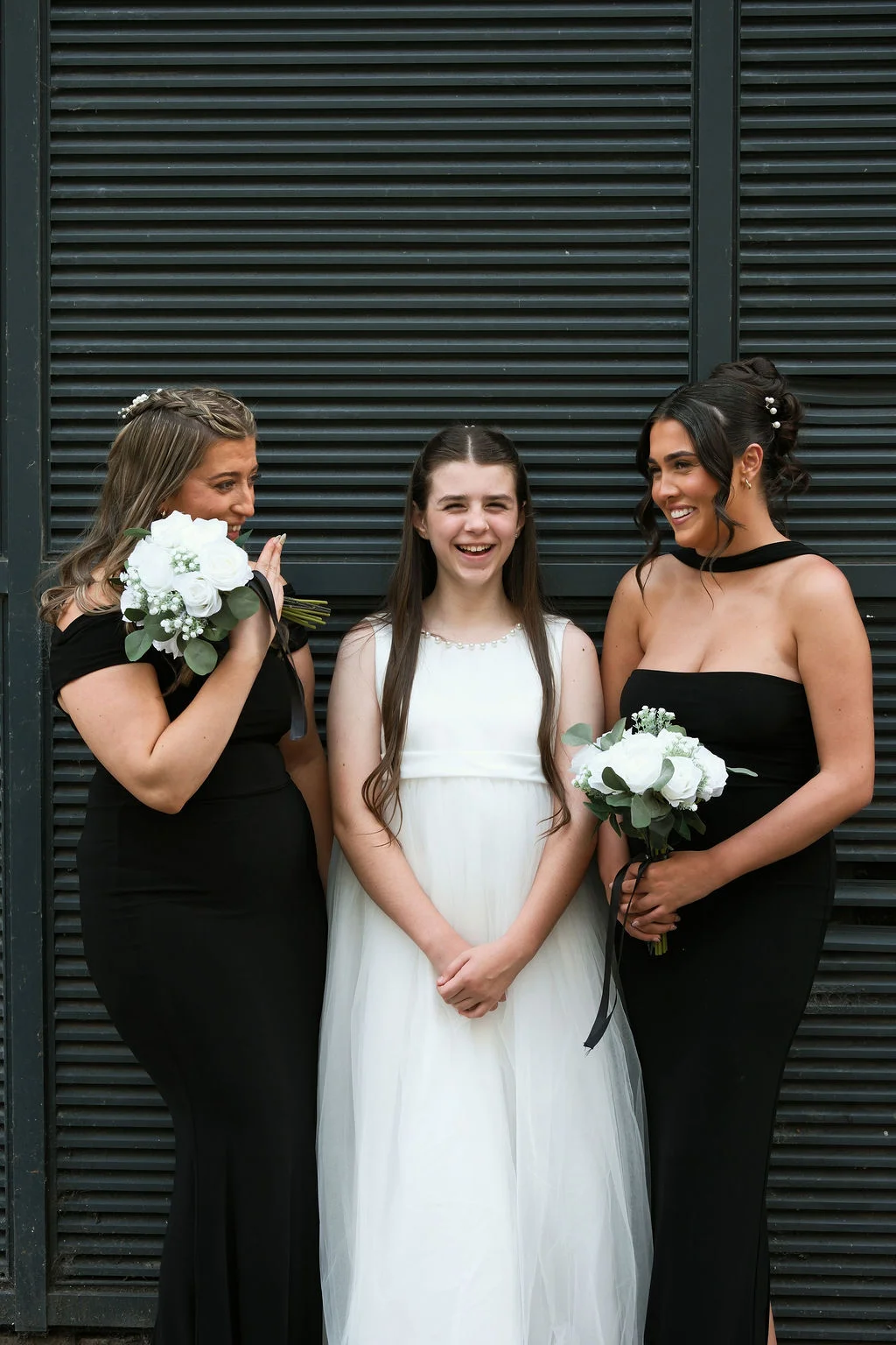 Two bridesmaids in elegant black gowns flank a junior bridesmaid in a white dress, all holding white rose bouquets and smiling against a dark slatted backdrop.