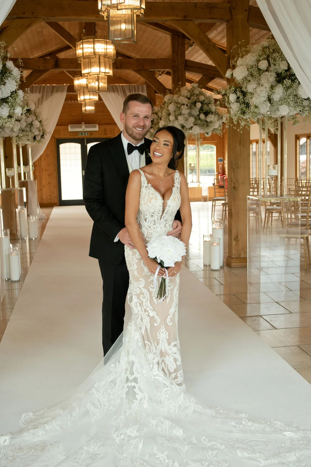 A smiling bride holding a white posy bouquet and groom in a black tuxedo embrace inside an oak-framed barn wedding venue.