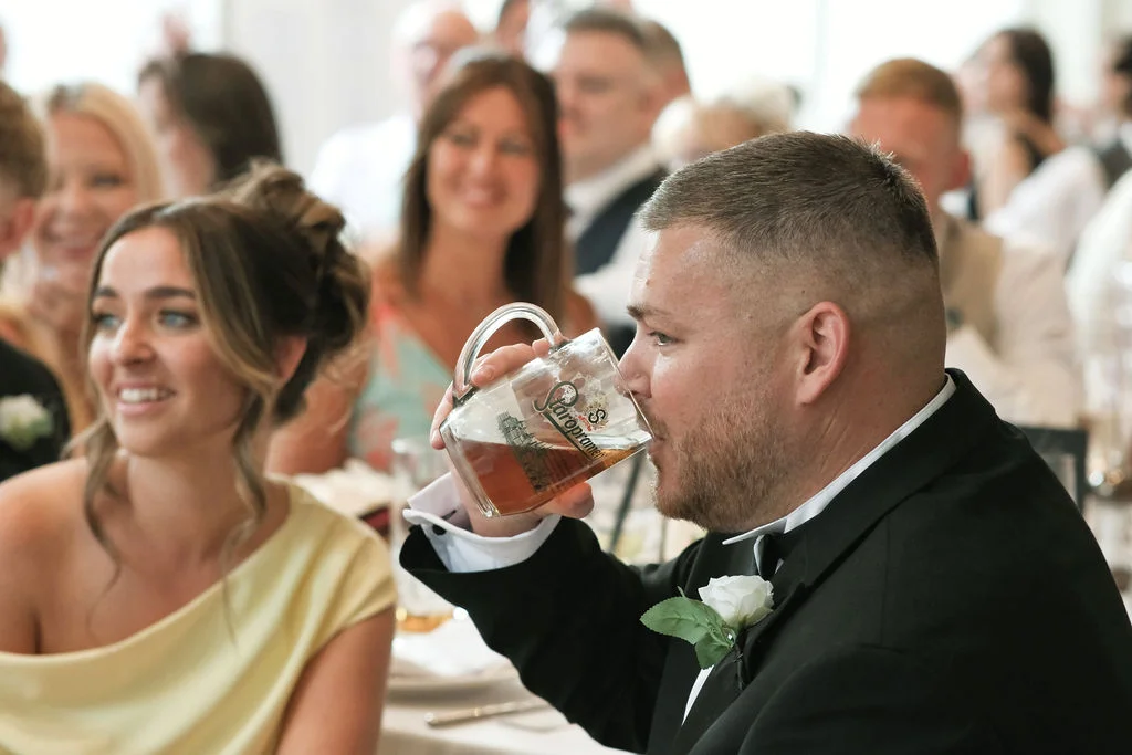A guest in a black tuxedo with a white buttonhole drinks from a beer glass during the wedding reception while other guests smile and watch nearby.