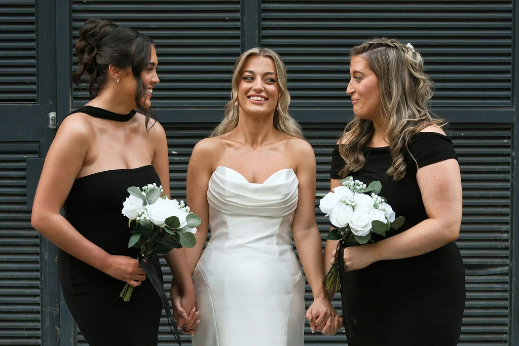 A smiling bride in a white strapless gown stands flanked by two bridesmaids in black dresses holding white bouquets against a dark slatted backdrop.
